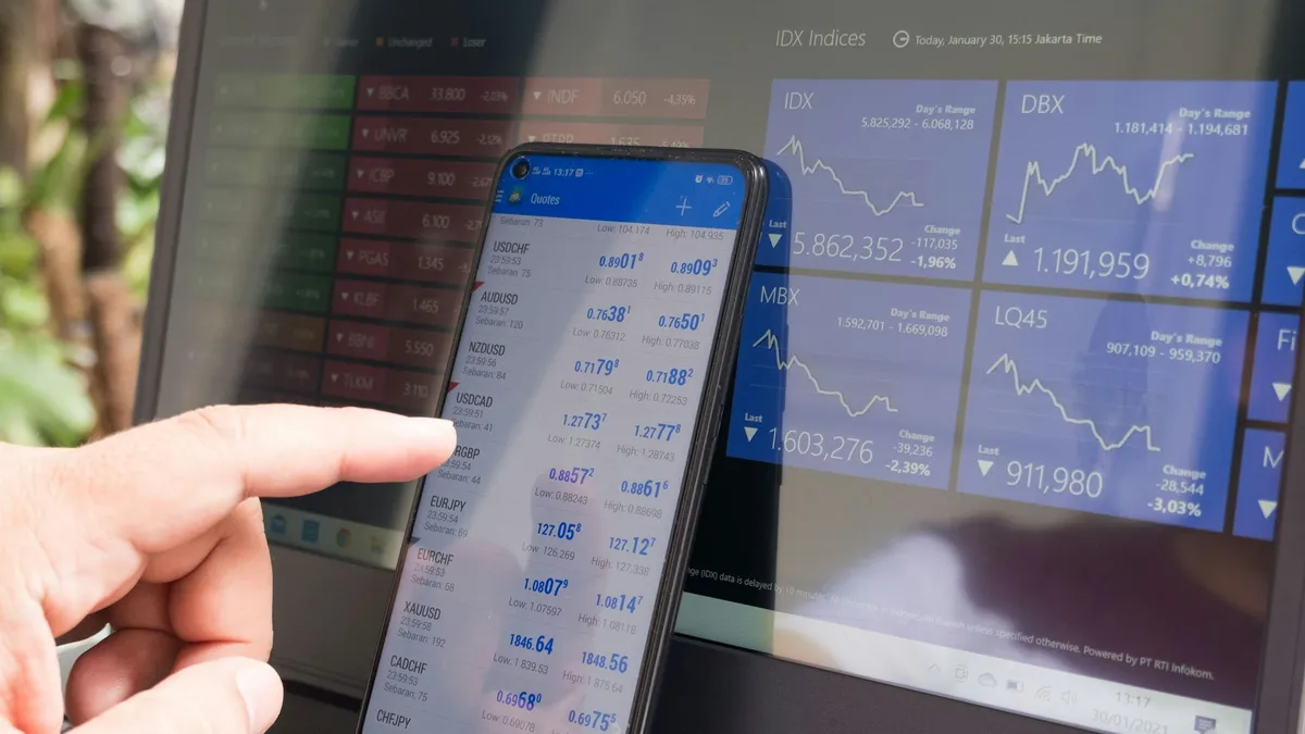 A hand pointing at a smartphone displaying currency quotes against a laptop.