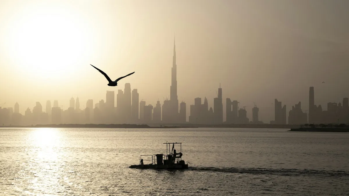 The Dubai skyline in haze across the water, with a gull in flight and a small boat in the foreground.