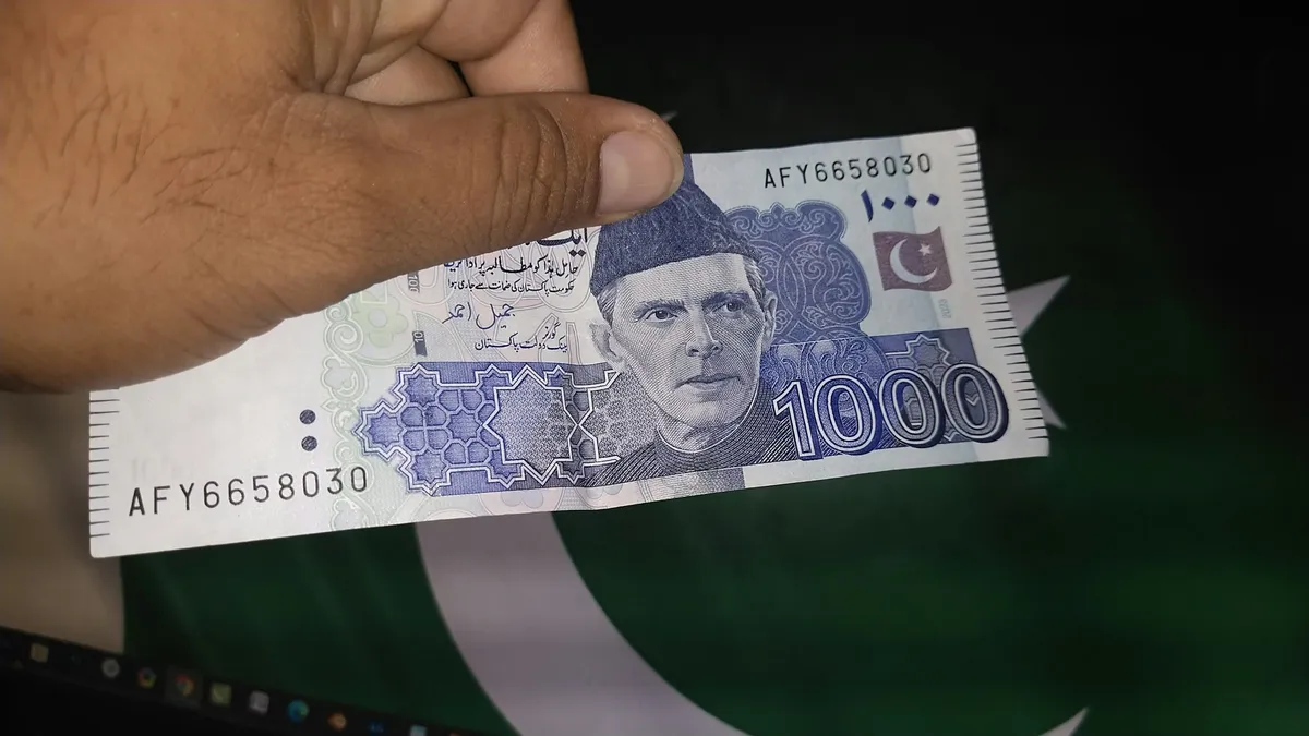 A hand holding a Pakistani Rs1,000 banknote in front of a Pakistan flag.