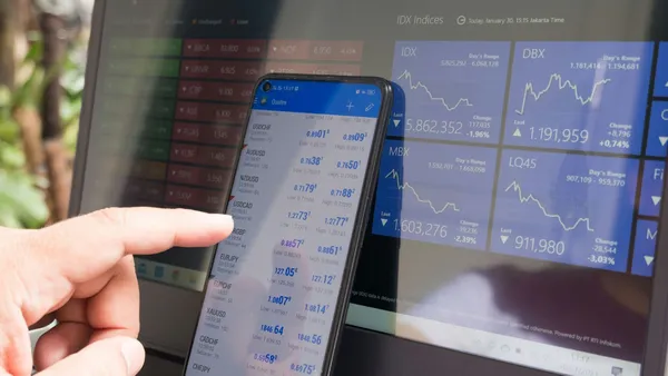 A hand pointing at a smartphone displaying currency quotes against a laptop.