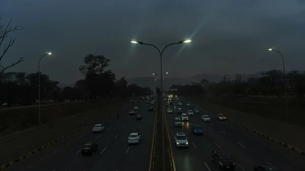 Evening traffic on a divided highway, street lights on, hills in the distance.